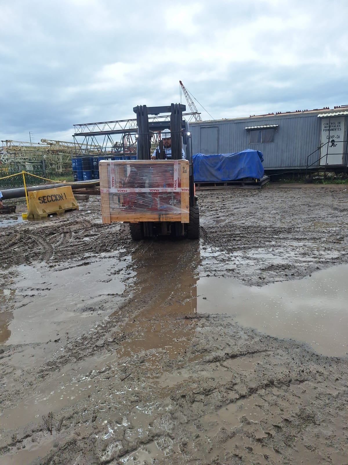 Montacargas moviendo una caja de madera marcada como frágil en un patio de materiales encharcado, con grúas de celosía al fondo