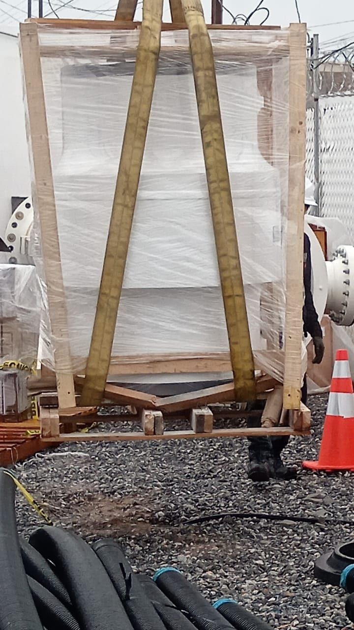 Equipo embalado en caja de madera y plástico, suspendido con eslingas sobre un terreno de grava en un sitio industrial