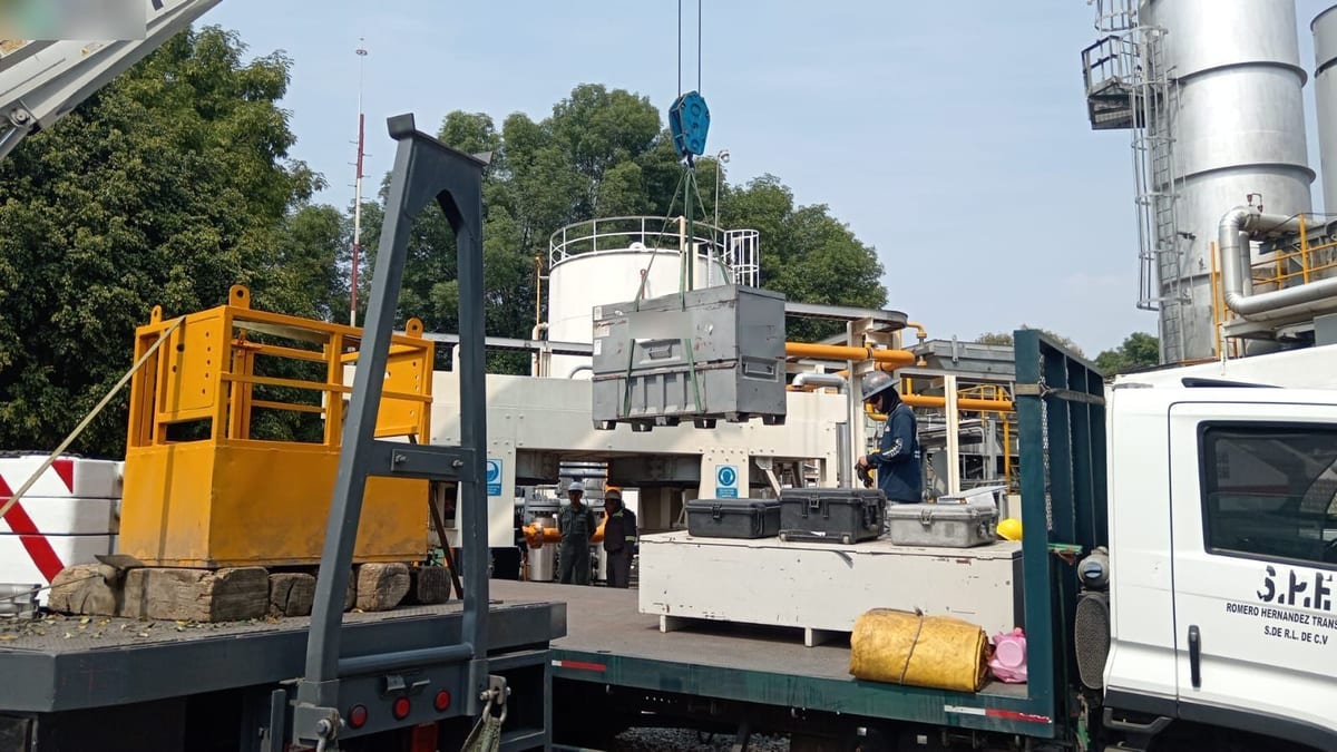 Maniobra de carga de equipo con grúa dentro de una planta industrial, con tanques y tubería al fondo